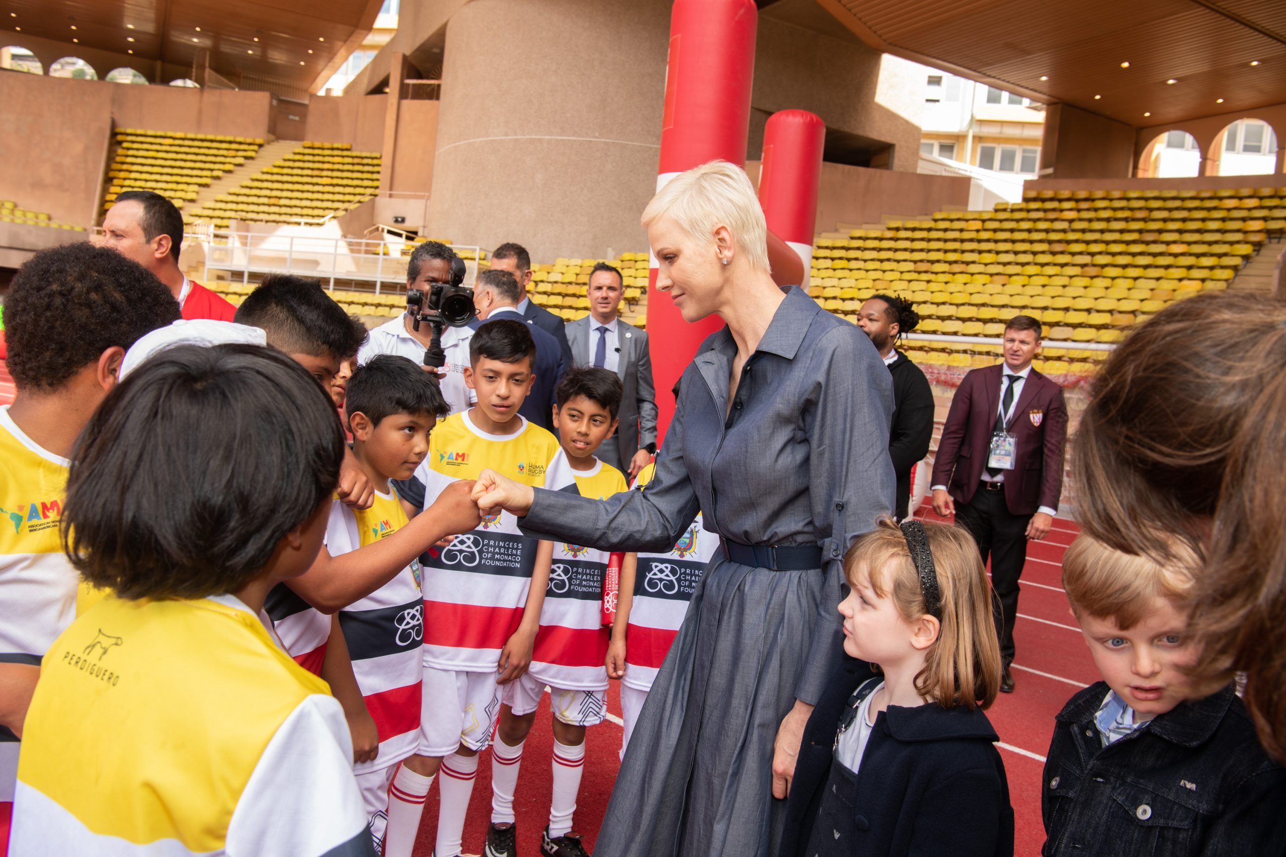 Una donna saluta con un pugno un gruppo di bambini in maglia sportiva, in uno stadio, durante un evento della Fondazione Princesse Charlène de Monaco