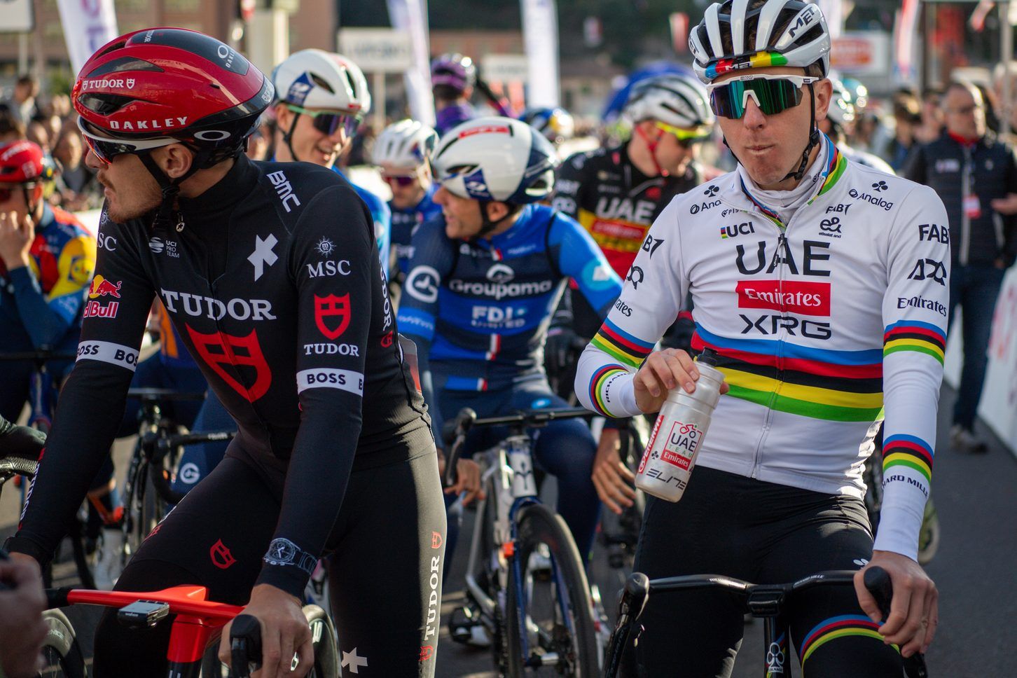 Due ciclisti in primo piano, uno in maglia nera Tudor e uno in maglia iridata UAE, con altri ciclisti sfocati dietro