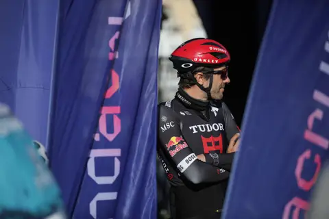 Ciclista in maglia nera Tudor e casco rosso, a braccia conserte tra bandiere blu, guarda di lato
