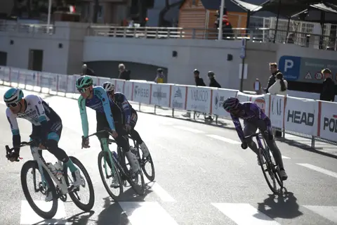 Quattro ciclisti in curva lungo le transenne, con lunghe ombre sull’asfalto e spettatori sullo sfondo