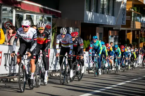 Gruppo di ciclisti in fila lungo le transenne, in testa maglie UAE, iridata e Tudor, con un hotel sullo sfondo