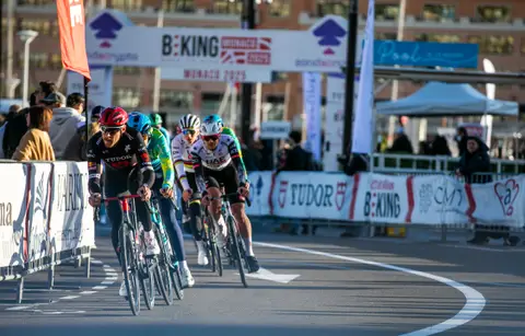 Gruppo di ciclisti in curva verso l’arco BEKING Monaco, con il pubblico dietro le transenne e un tendone bianco