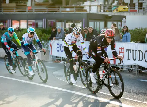 Ciclisti in curva lungo le transenne Tudor, uno in maglia iridata, con spettatori sullo sfondo e luce radente