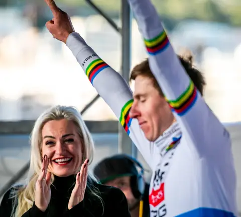 Ciclista in maglia iridata con le braccia alzate; accanto, una donna bionda applaude sorridendo