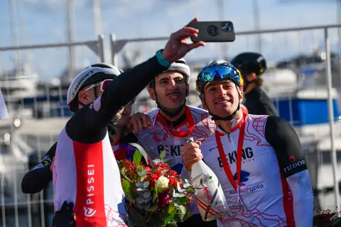 Tre ciclisti in maglia da squadra posano per un selfie con un mazzo di fiori e una bottiglia, con il porto dietro