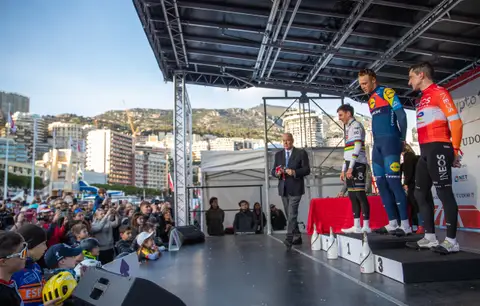 Premiazione sul palco con tre ciclisti e un uomo in giacca e cravatta, il pubblico a sinistra e le colline dietro