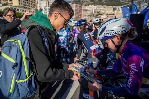 Ciclista in maglia viola e casco bianco firma un opuscolo a una persona con zaino e occhiali