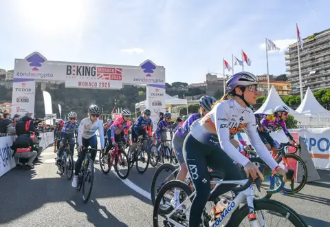 Gruppo di ciclisti sotto l’arco BEKING Monaco; in primo piano una maglia UAE bianca e un casco viola