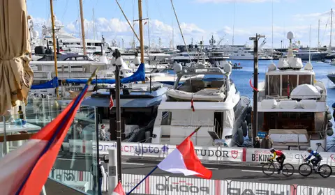 Due ciclisti passano lungo le transenne del porto di Monaco, con yacht ormeggiati e bandiere in primo piano