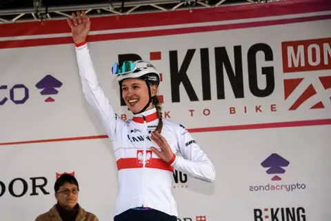 Ciclista sorridente con la treccia, in maglia bianca e rossa, casco bianco e un braccio alzato, davanti al fondale BEKING