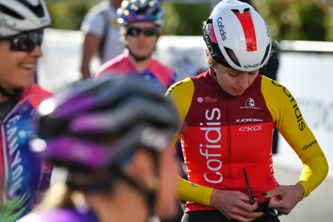 Cyclist in a red and yellow Cofidis jersey and white helmet looks down, sunglasses in hand; blurred purple helmet in front