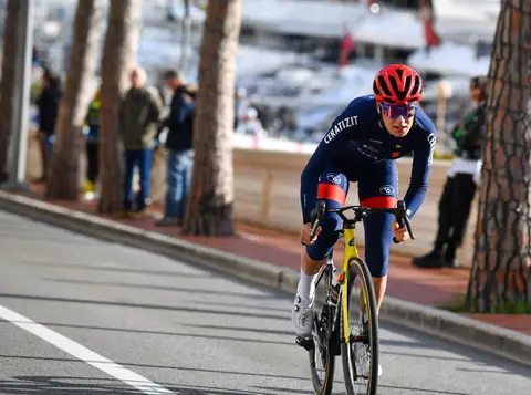 Ciclista in tenuta blu Ceratizit e casco rosso pedala su una bici gialla, con alberi e spettatori sullo sfondo