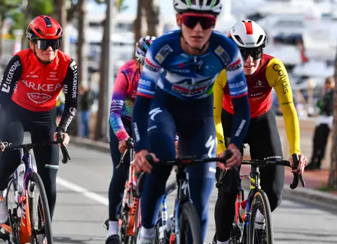 Gruppetto di almeno cinque ciclisti ripreso di fronte su un viale alberato, in maglie Arkéa, Cofidis e blu, porto dietro