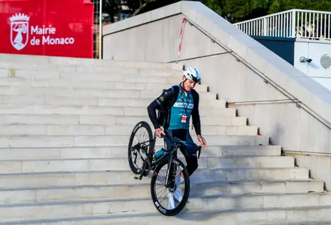 Ciclista con casco bianco scende una scalinata chiara con la bici sollevata, sotto uno striscione Mairie de Monaco