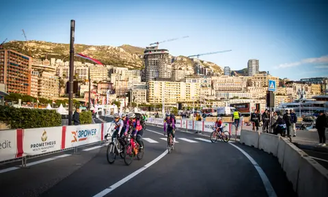 Piccolo gruppo di ciclisti in curva lungo le transenne, con palazzi e colline di Monaco sullo sfondo e cielo sereno