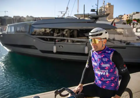 Ciclista in maglia viola a maniche nere e casco bianco, appoggiato alla bici sul molo davanti a uno yacht