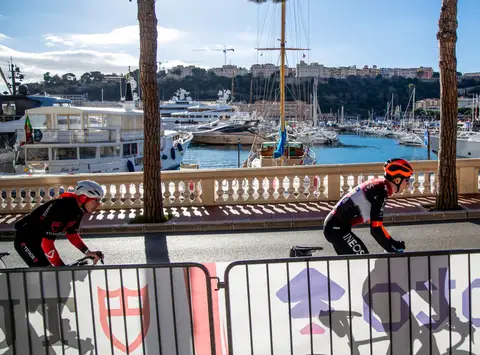 Due ciclisti in maglia Tudor e Ineos pedalano dietro le transenne, con balaustra, porto e barche sullo sfondo