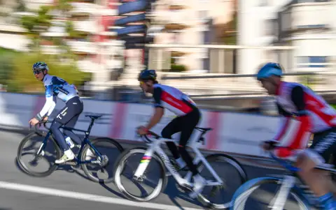 Tre ciclisti ripresi in panning su strada, con edifici e transenne sfocati dal movimento sullo sfondo