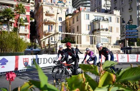 Gruppo di ciclisti pedala davanti alle transenne Tudor e ai palazzi, con foglie verdi in primo piano
