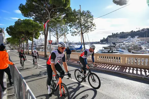 Due ciclisti in maglia BEKING in primo piano lungo la balaustra del porto, altri due dietro, sotto i pini