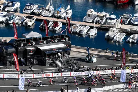 Veduta dall’alto dello stand nero Tudor sul porto, con bandiere rosse, barche ormeggiate e un gruppo di ciclisti in corsa