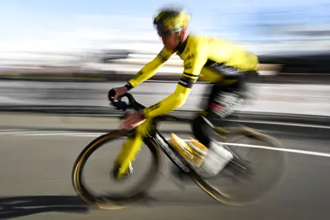 Ciclista in maglia e casco gialli, di profilo, ripreso in panning con sfondo e strada sfocati dal movimento