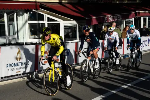 Quattro ciclisti in fila lungo le transenne, il primo in maglia gialla e nera, con spettatori e tende rosse dietro