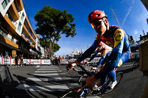 Ciclista in maglia Lidl e casco rosso in primo piano, ripresa fisheye; dietro un altro ciclista, il porto e i pini