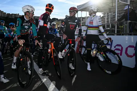 Quattro ciclisti fermi sulle bici, in maglia Tudor e UAE, parlano fra loro; altri ciclisti e una tenda bianca dietro