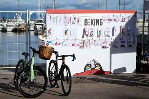 Pannello BEKING con disegni di biciclette appesi con mollette e due bici parcheggiate davanti, sul molo