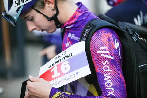 Ciclista in maglia viola, casco bianco e zaino nero guarda in basso il pettorale numero 16 che regge davanti al corpo