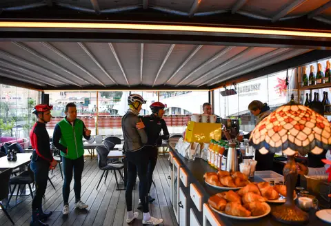 Tre ciclisti con il casco e un uomo in gilet verde vicino al bancone della colazione con cornetti e succhi