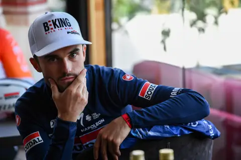 Ciclista con cappellino BEKING e maglia blu Soudal, il mento appoggiato alla mano, davanti a una vetrata