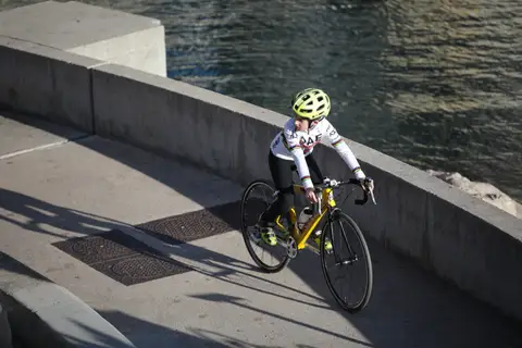 Giovane ciclista con casco giallo fluo e maglia bianca a bande arcobaleno pedala lungo un muretto, vista dall’alto