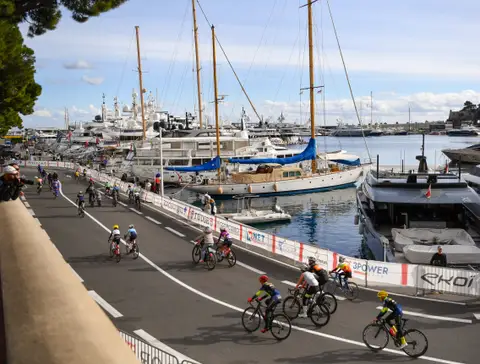 Ciclisti su un tratto di strada lungo il porto, con yacht e barche a vela ormeggiati oltre le transenne