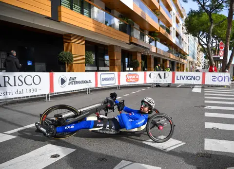 Atleta in handbike in tuta azzurra Italia percorre la strada davanti a transenne con banner di sponsor
