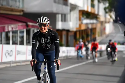 Ciclista in maglia nera UAE, casco e occhiali a specchio, sorride in sella; dietro altri ciclisti sfocati