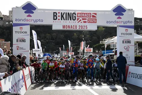 Giovani ciclisti schierati al via sotto l’arco BEKING Monaco, con gli spettatori dietro le transenne