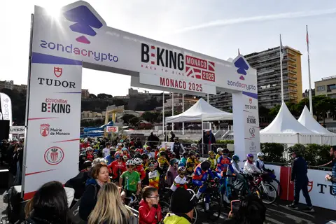 Giovani ciclisti schierati sotto l’arco BEKING Monaco, ripresi in controluce, con spettatori in primo piano