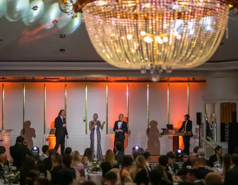 Tre persone sul palco della cena di gala e un DJ alla consolle, sotto un grande lampadario di cristallo