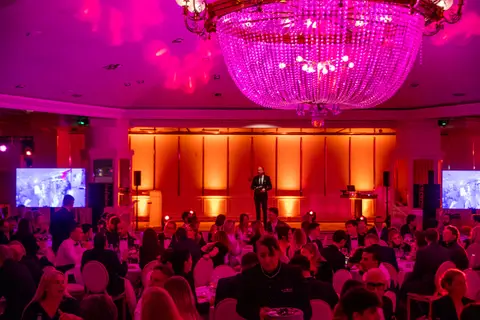 Sala della cena di gala con luci rosa e lampadario di cristallo: un uomo sul palco, ospiti ai tavoli e due schermi