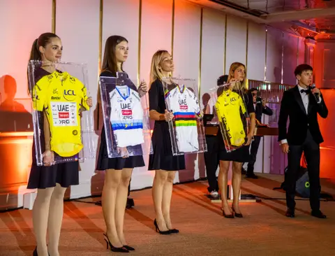 Quattro donne in nero reggono maglie incorniciate, due gialle e due bianche; a destra un uomo in smoking