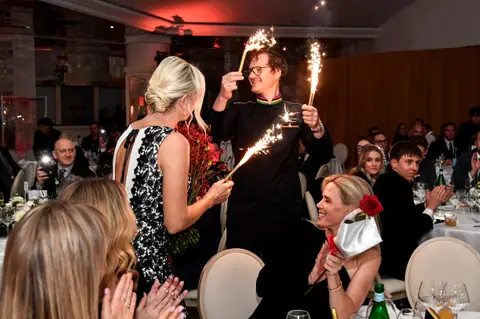 Uomo con due candele scintillanti tra i tavoli della cena di gala, tra applausi e una donna con fiori