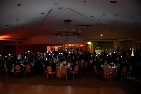 Sala della cena di gala al buio, con gli ospiti ai tavoli rotondi che alzano telefoni con la torcia accesa