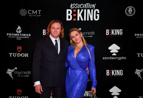 Un uomo in completo scuro e una donna in abito blu glitterato posano davanti al fondale Association BEKING