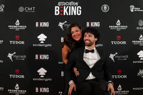 Uomo in smoking e papillon su una sedia a rotelle sorride con una donna alle sue spalle, davanti al fondale BEKING